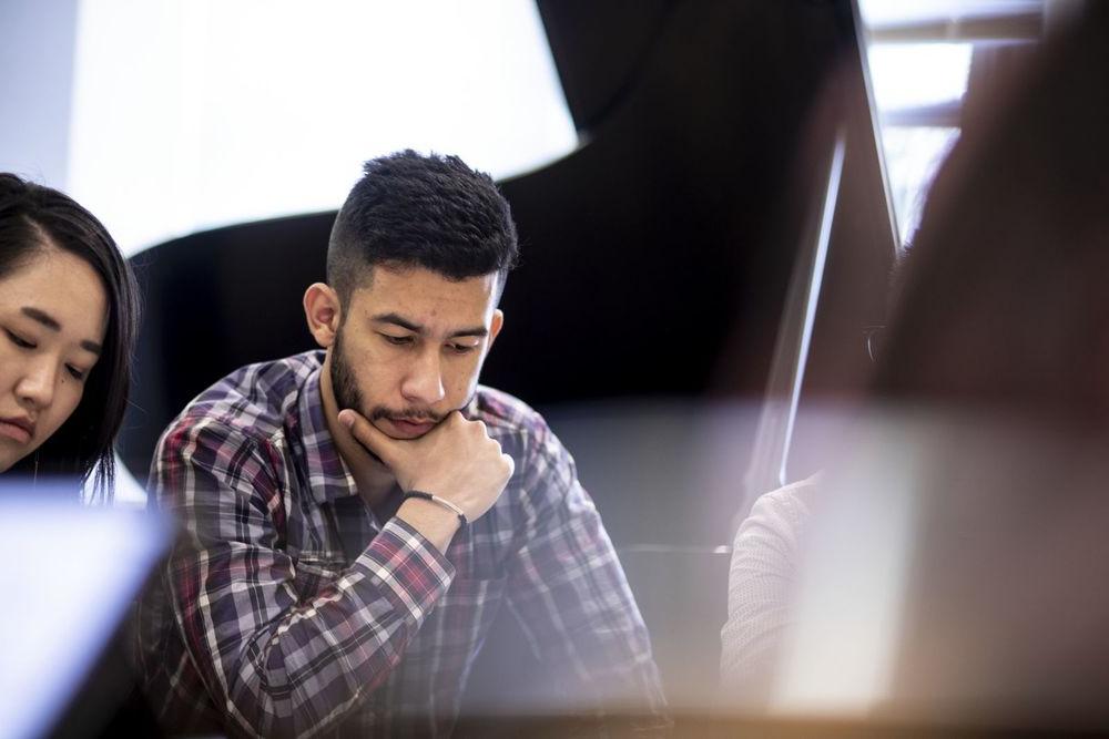 A Music Theory student, with hand on chin, looks down at his work while sitting among other students.